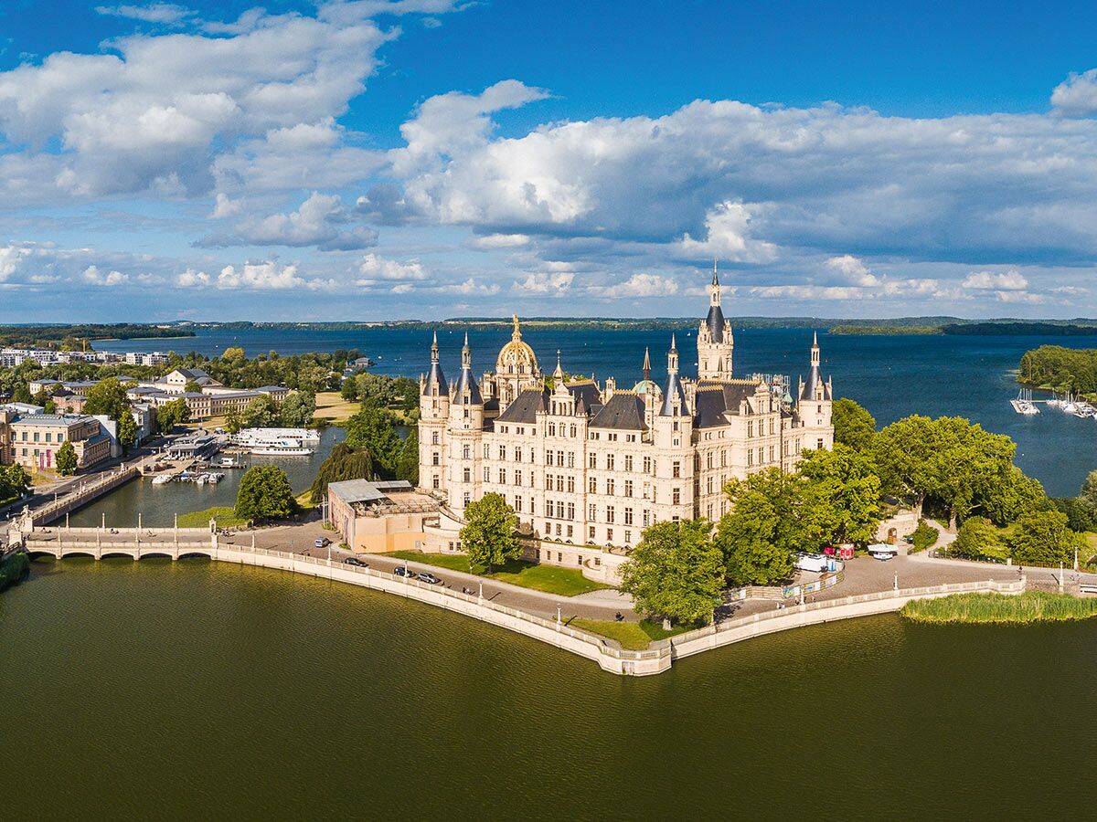 Majestätisches Schweriner Schloss, von Wasser umgeben, mit Brücken und grüner Landschaft unter blauem Himmel und Wolken.
