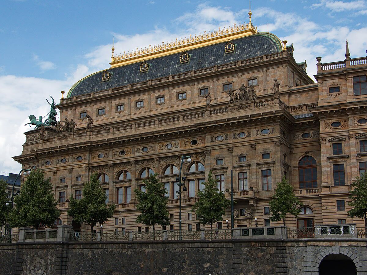 Blick auf das prächtige Nationaltheater in Prag mit goldener Kuppel, Skulpturen und steinerner Fassade am Flussufer.