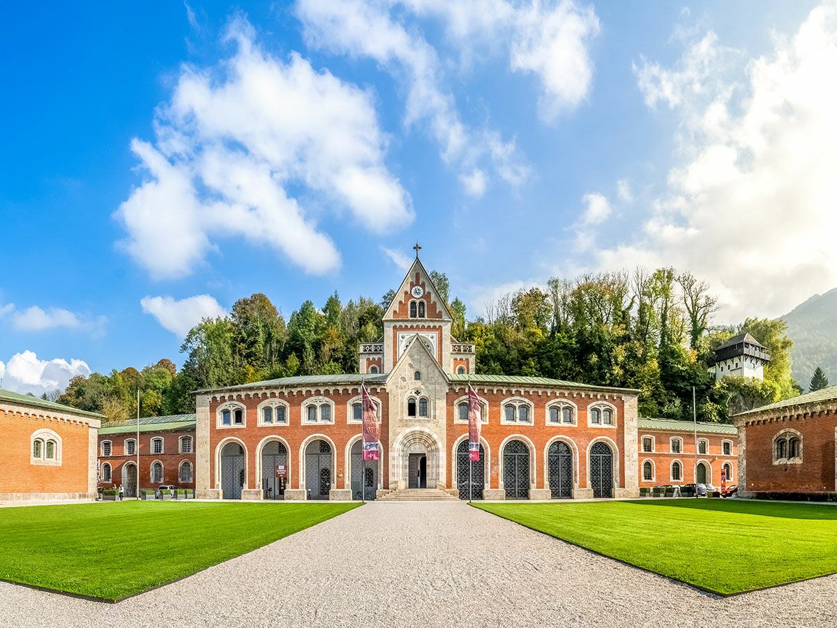 Die historische Winterreitschule in Salzburg mit rotem Backstein, Bögen, Uhrturm und grünen Wiesen, umgeben von Bergen unter blauem Himmel.