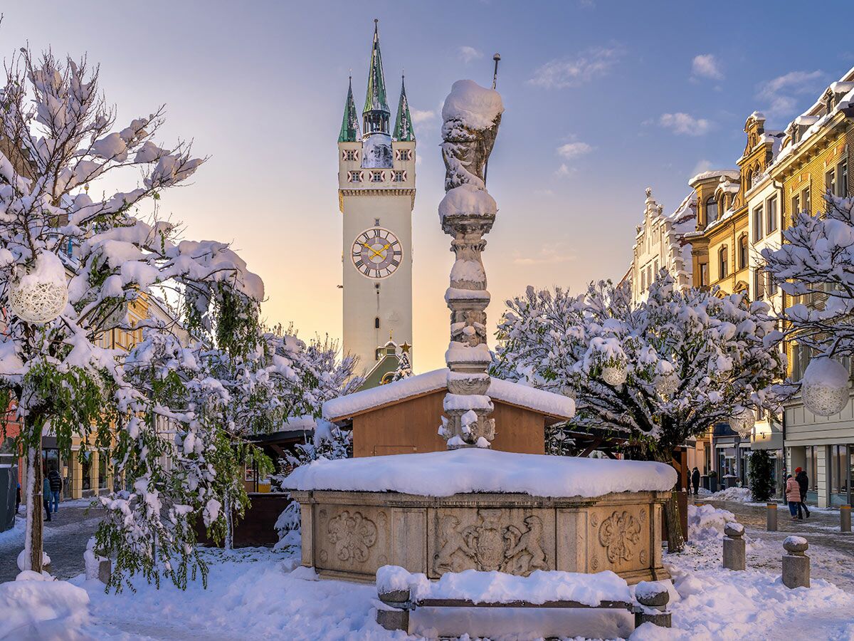 Malerischer verschneiter Marktplatz in Baden-Baden mit historischem Stadtturm, Steinbrunnen und schneebedeckten Bäumen bei Sonnenuntergang.