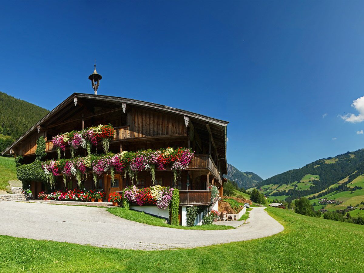 Idyllisches Alpenhaus in den Bergen mit üppig blühenden Blumenbalkonen, grünem Gras und blauem Himmel bei Sonnenschein.
