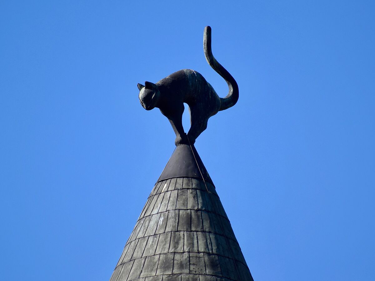Eine schwarze Katzenskulptur mit erhobenem Schwanz thront auf einem dunklen, kegelförmigen Dach vor blauem Himmel.