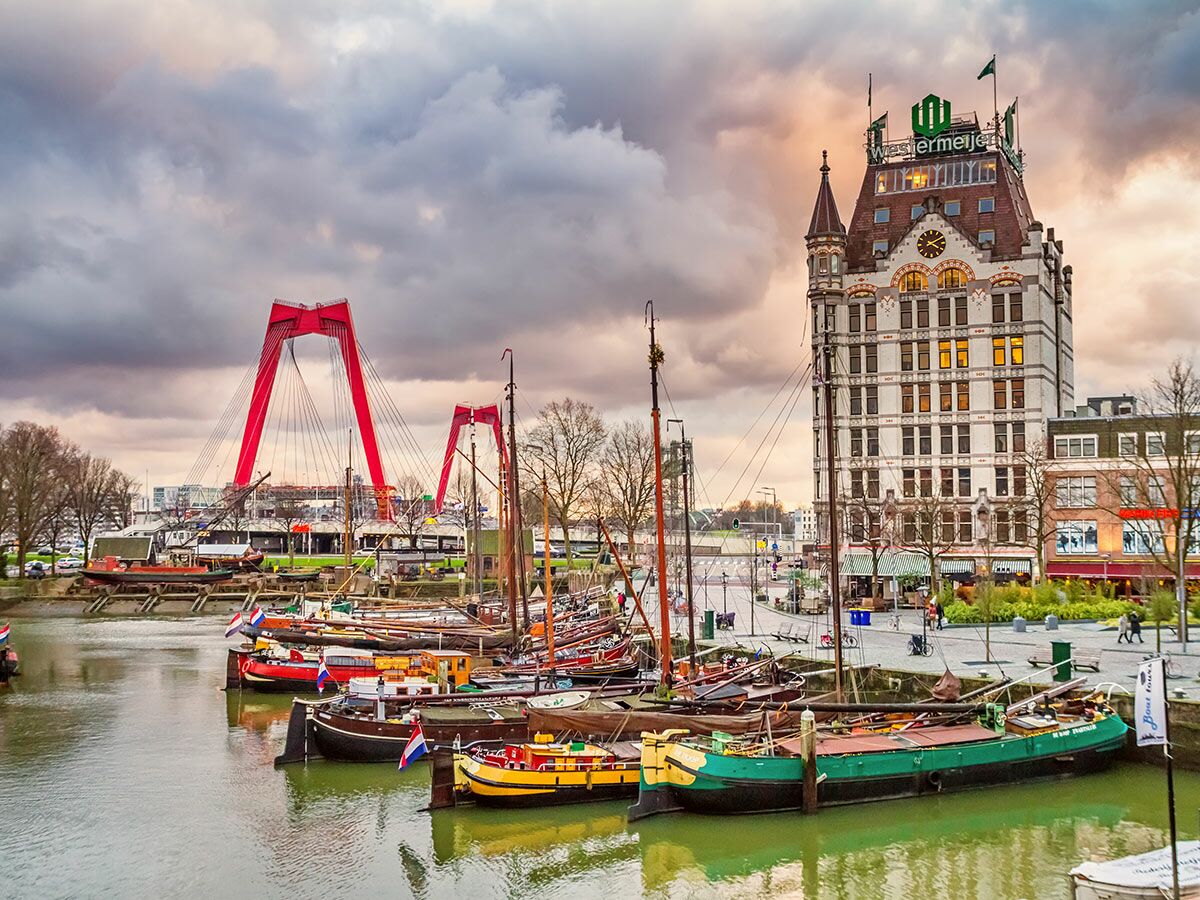 Panoramablick auf Rotterdams Oude Haven mit historischen Booten, dem Weißen Haus und der roten Erasmusbrücke bei Sonnenuntergang.