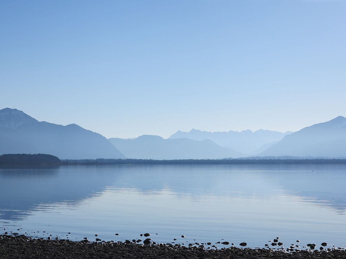 Ruhiger Bergsee mit spiegelnden blauen, in Dunst gehüllten Bergen im Hintergrund unter klarem Himmel.