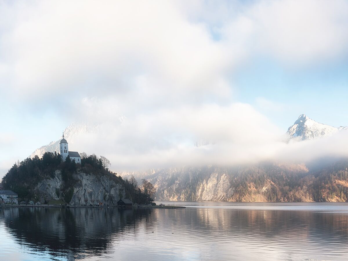 Johannesbergkapelle auf felsiger Halbinsel im Traunsee, umgeben von Nebel und verschneiten Bergen unter blauem Himmel.