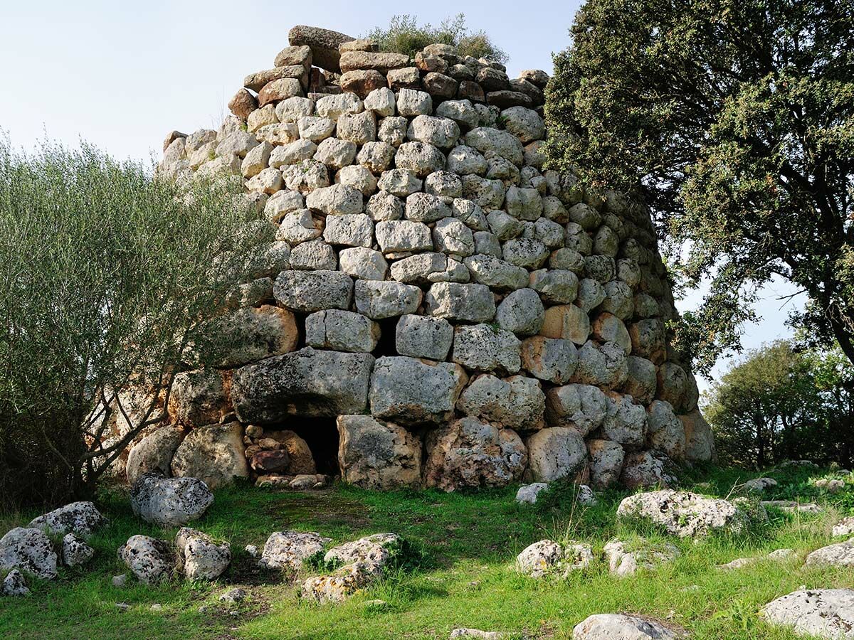 Prähistorischer Nuraghe-Steinturm in Sardinien mit Eingang, umgeben von Bäumen und grünem Gras unter hellem Himmel.
