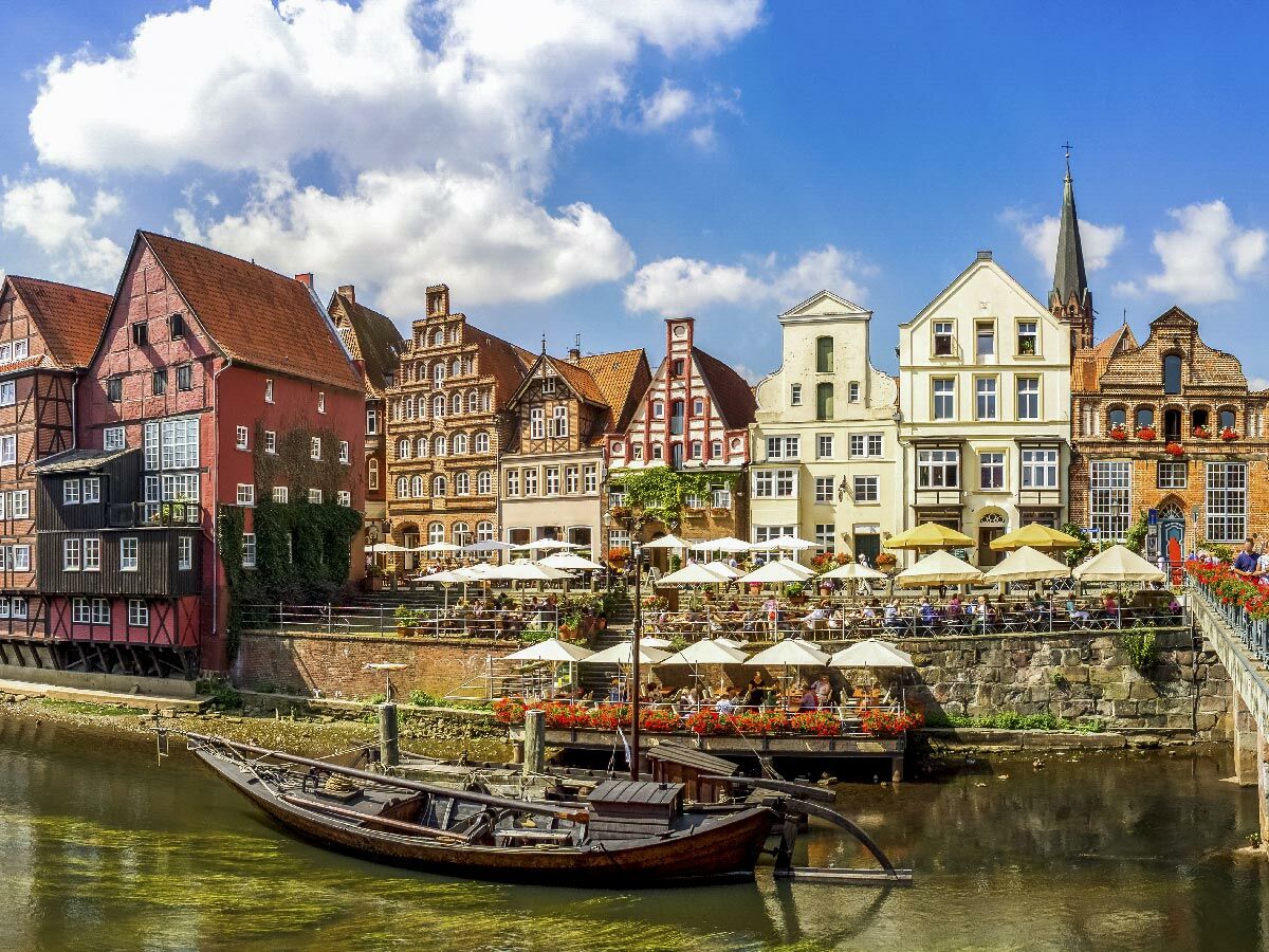 Reihe historischer Fachwerkhäuser und Giebelhäuser an einem Flussufer mit belebten Straßencafés und einem Holzboot im Vordergrund.