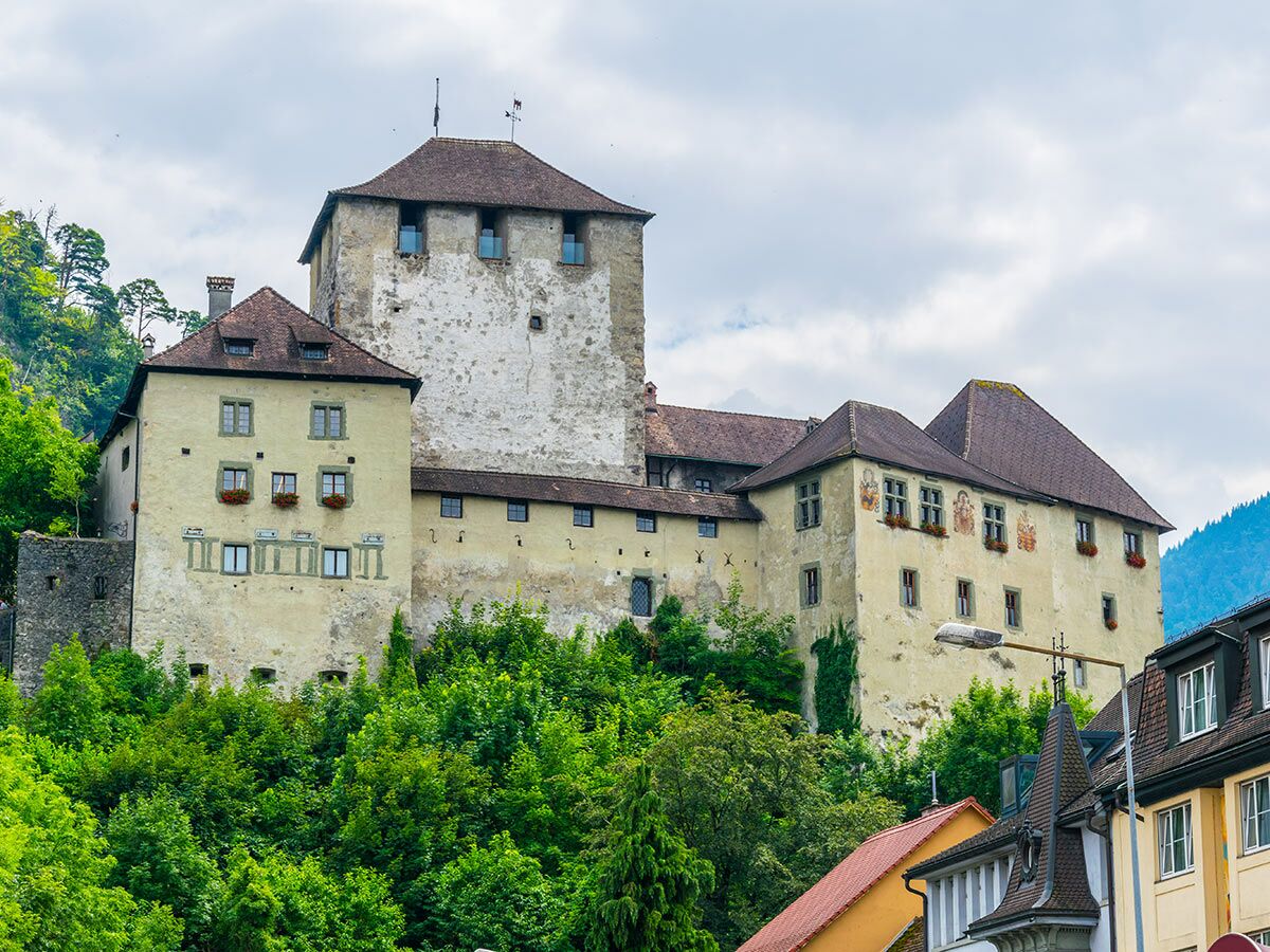 Historisches Schloss Landeck in Tirol, Österreich, auf einem Hügel umgeben von grünen Bäumen unter bewölktem Himmel.