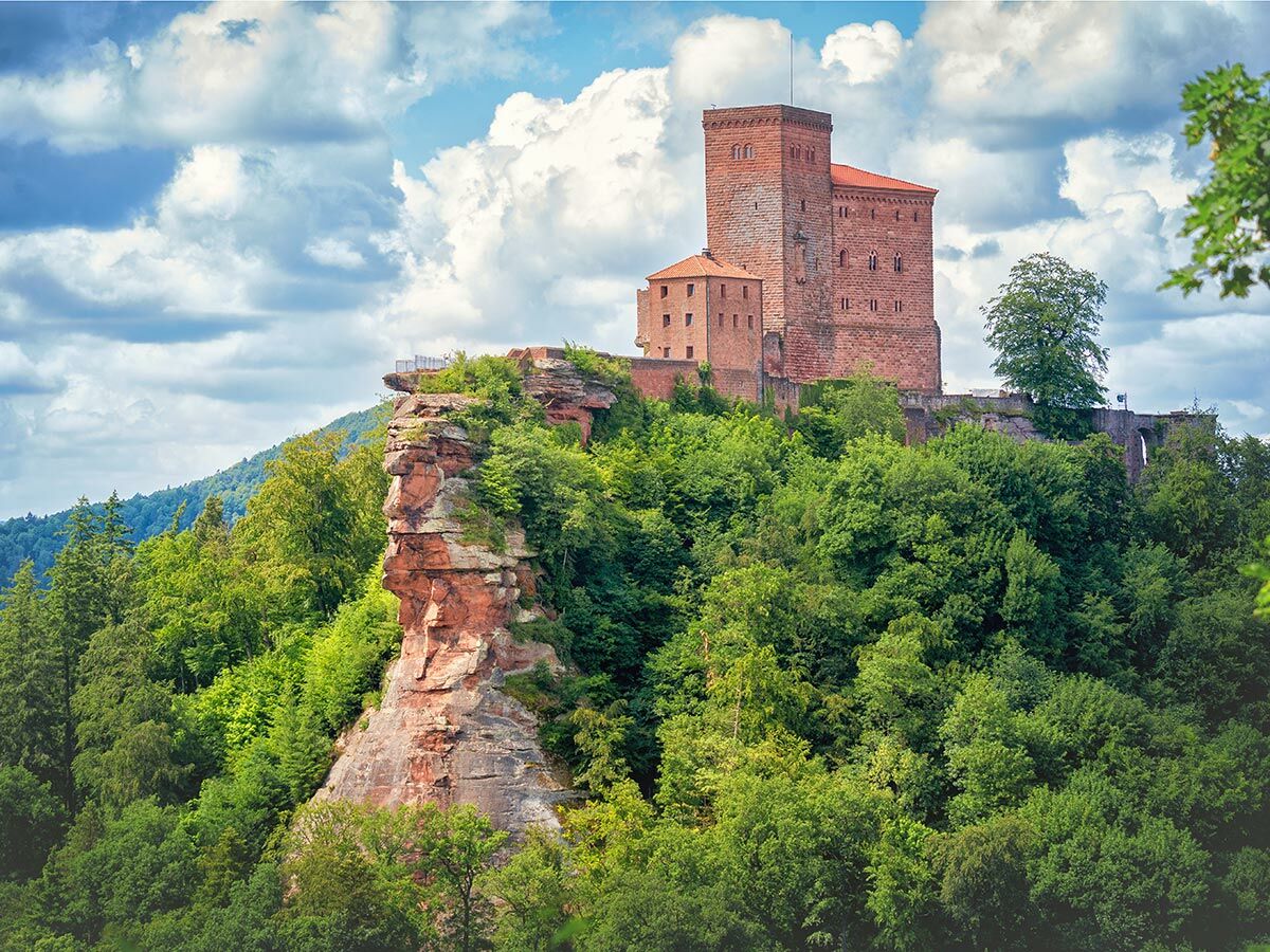Historische Burg aus rotem Sandstein thront auf einem bewaldeten Felsen unter blauem Himmel mit weißen Wolken.
