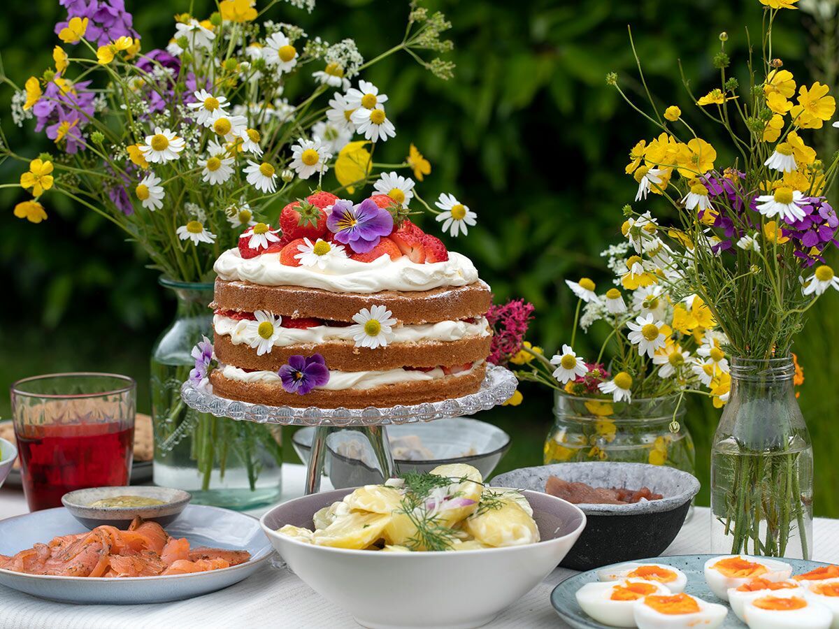Festlich gedeckter Tisch im Garten mit Erdbeerkuchen, Salaten, Eiern, Lachs und Wildblumen-Bouquets.