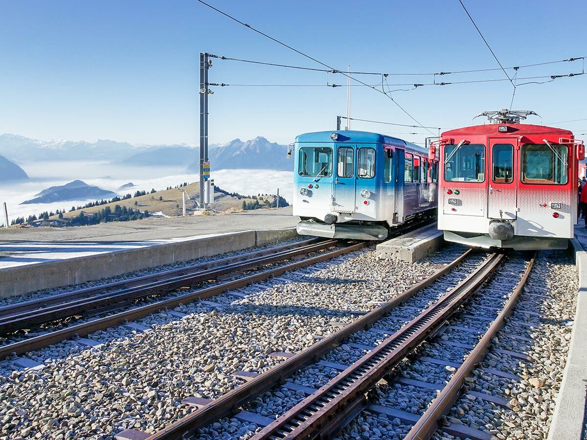 Zwei bunte Rigi-Bergbahnen (blau, rot) an einem Bahnhof in den Alpen, mit Nebelmeer und Gipfeln im Hintergrund.