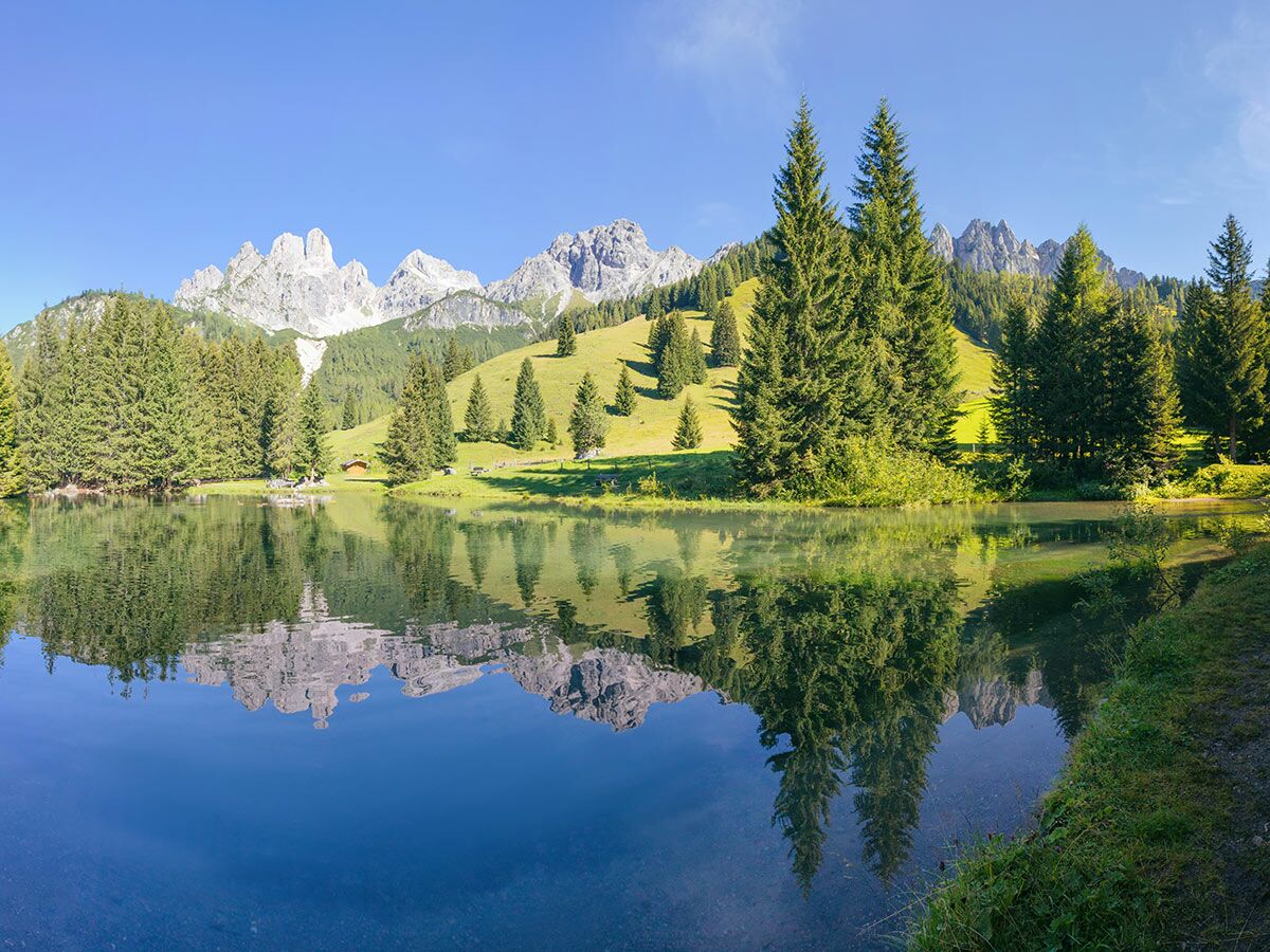 Idyllischer Bergsee in den Alpen mit kristallklarer Spiegelung von majestätischen Bergen, grünen Wäldern und blauem Himmel.