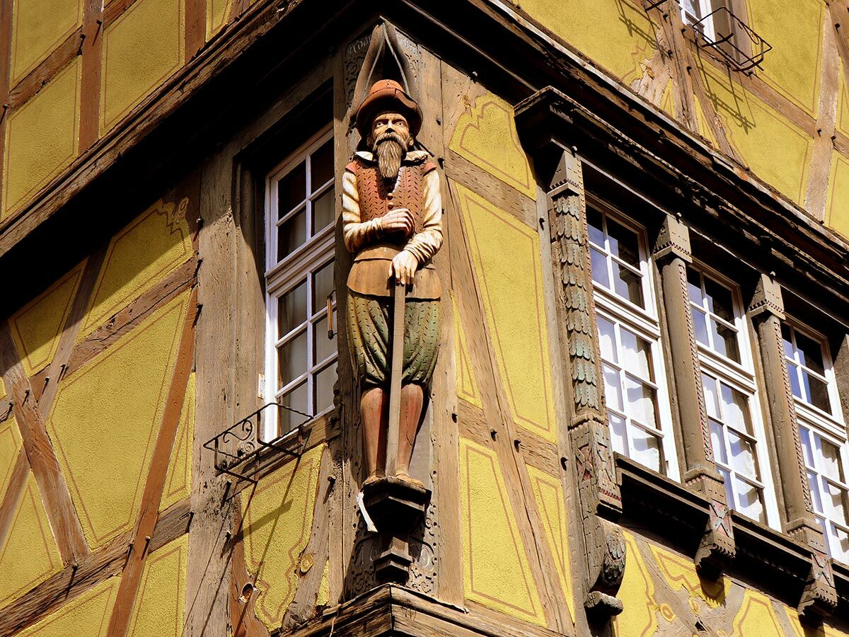 Detaillierte Holzstatue eines bärtigen Mannes an der Ecke eines gelben Fachwerkhauses mit Fenstern.