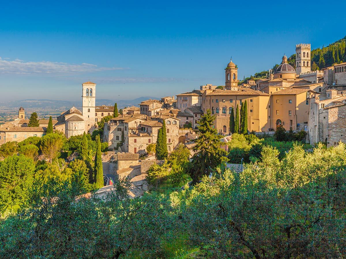 Malerische historische Stadt Assisi in Umbrien, Italien, mit alten Gebäuden, Kirchen und grünen Bäumen unter blauem Himmel.