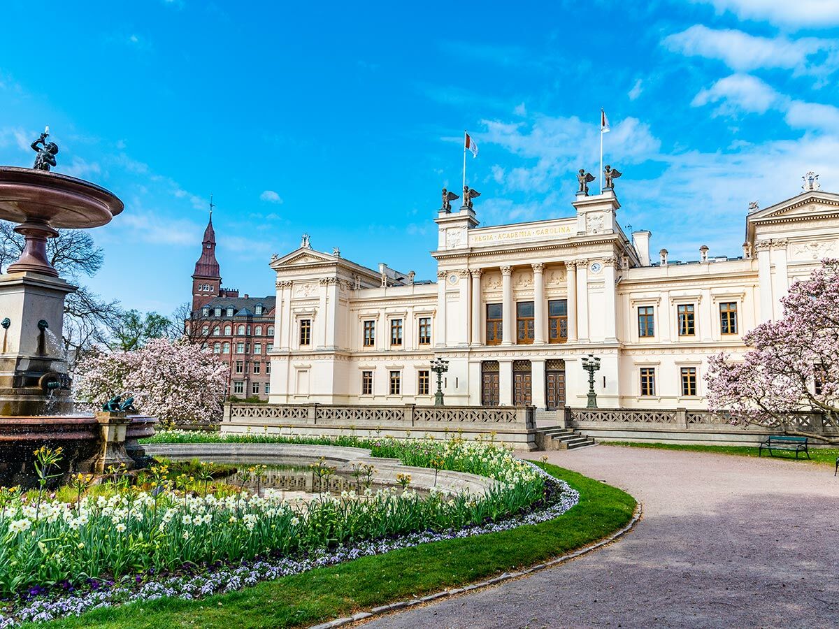 Das historische Hauptgebäude der Universität Lund in Schweden im Frühling, mit Brunnen, rosa Magnolien und Blumenbeeten unter blauem Himmel.