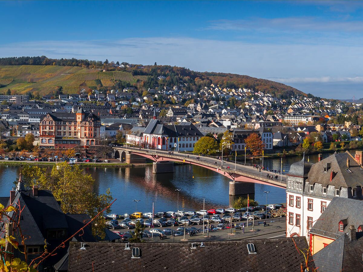 Panorama von Bernkastel-Kues an der Mosel mit historischer Stadt, Brücke, Herbstlaub und Weinbergen unter blauem Himmel.