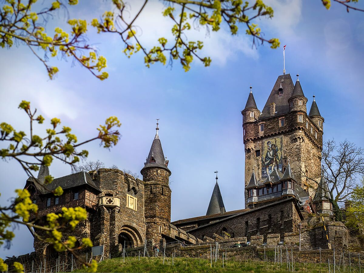 Historische Reichsburg Cochem an der Mosel mit Türmen, Steinmauern und einem Wandgemälde, umrahmt von frühlingshaften Ästen.
