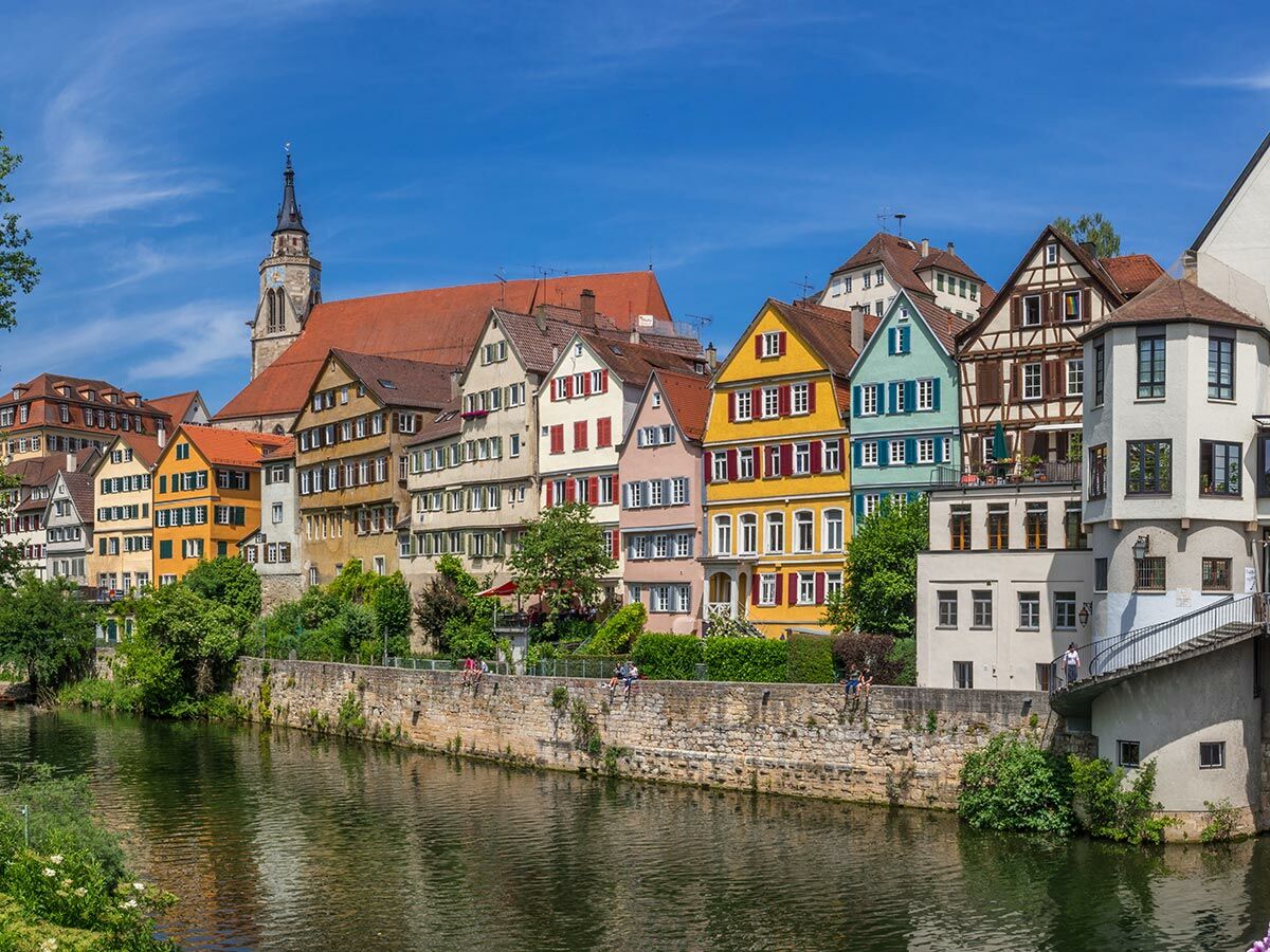 Bunte Häuser der Neckarfront in Tübingen am Fluss Neckar unter blauem Himmel mit Menschen auf der Mauer.