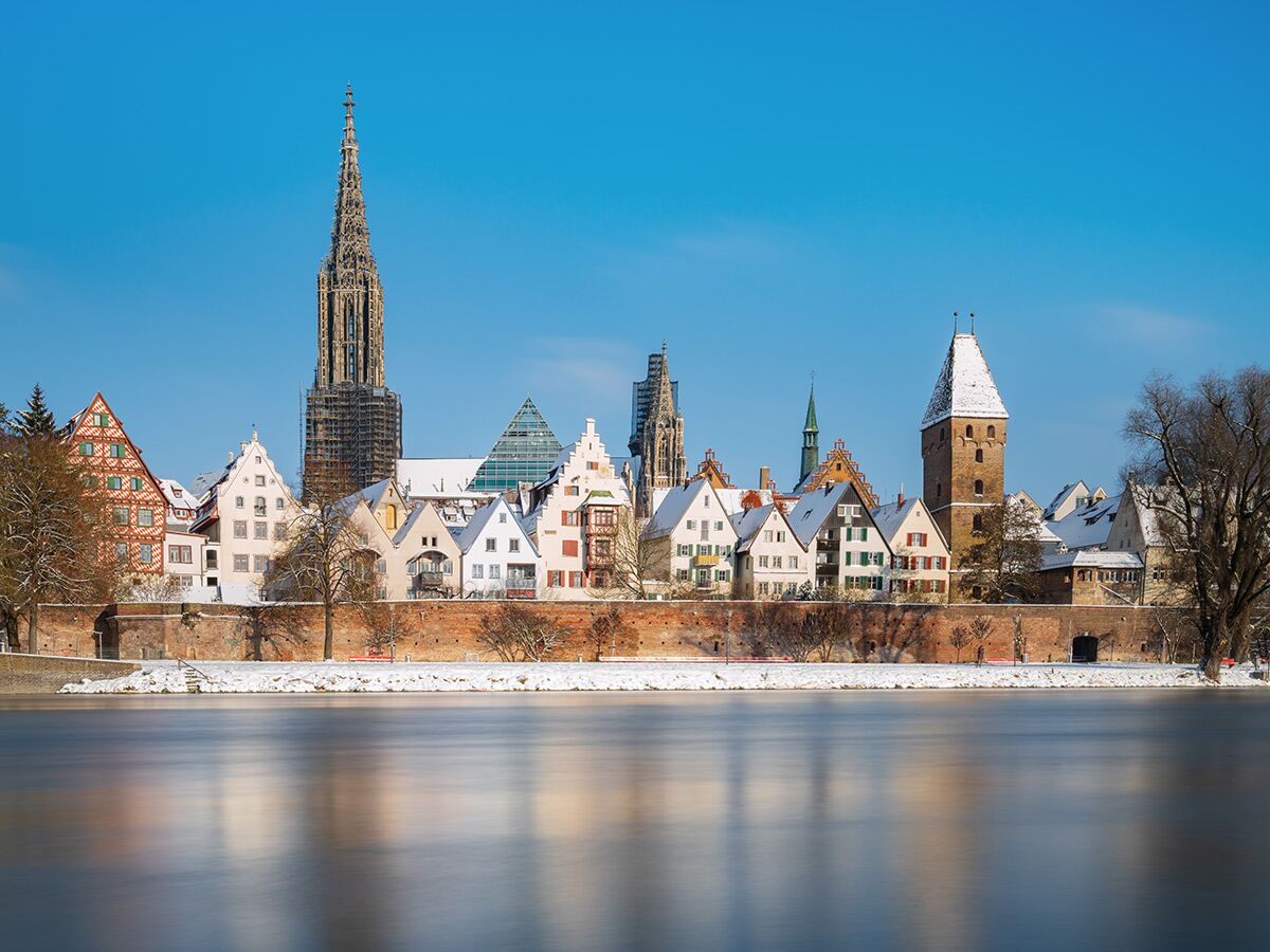 Winterlandschaft Ulm mit verschneiten historischen Gebäuden, Münster, Metzgerturm und Donau bei blauem Himmel.