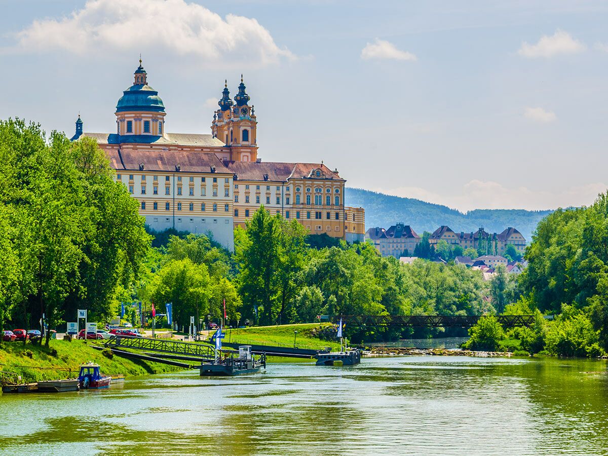 Prächtiges Stift Melk mit barocker Architektur am Flussufer der Donau in Österreich, umgeben von grünen Bäumen an einem sonnigen Tag.