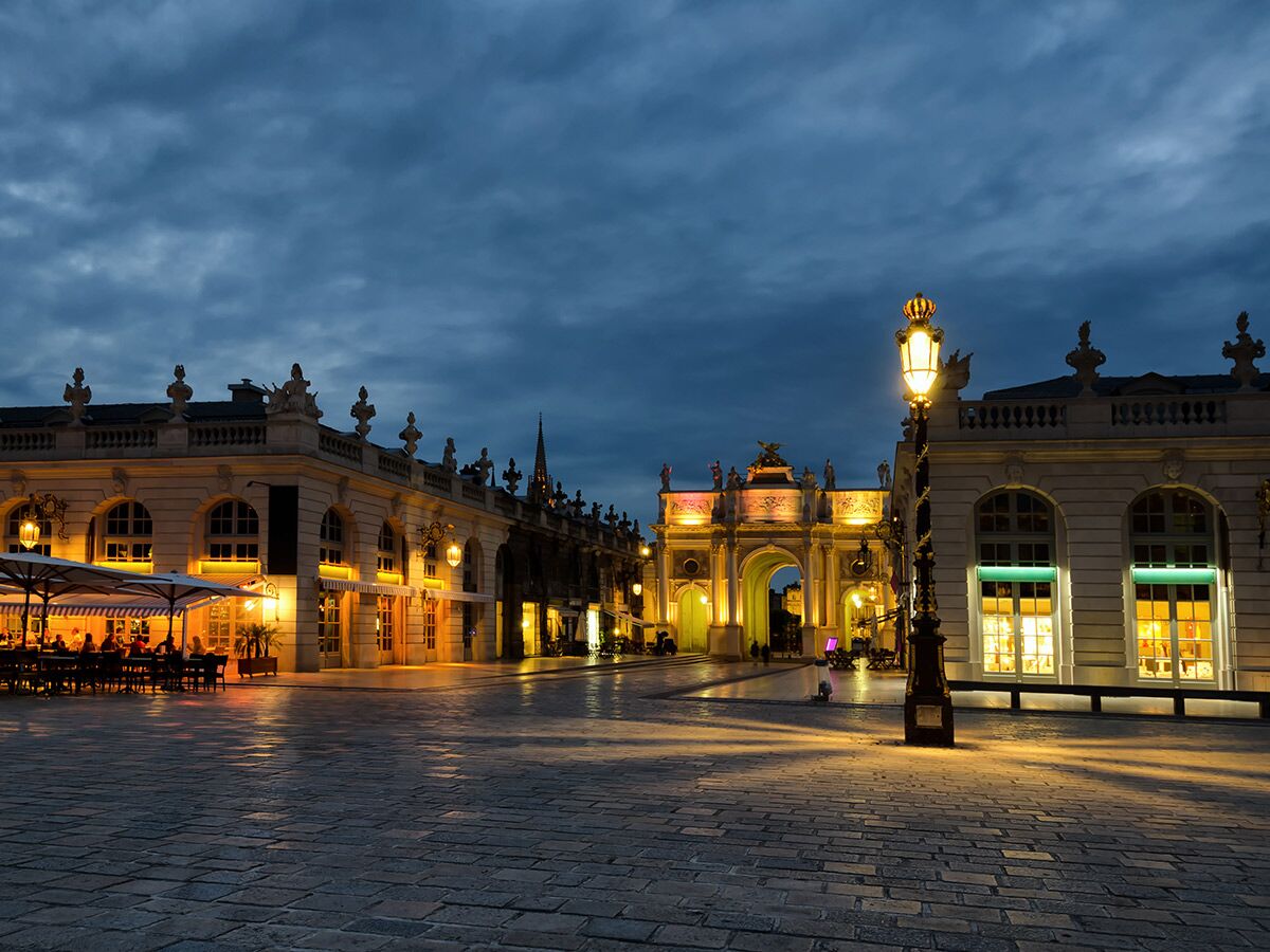 Abendansicht des beleuchteten Place Stanislas in Nancy mit historischen Gebäuden, Triumphbogen und Restaurants.