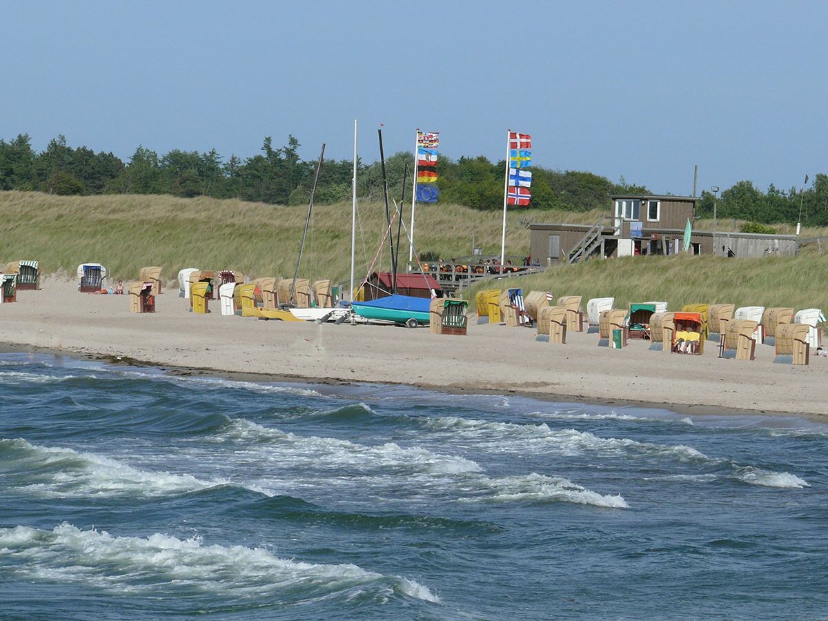 Bunter Strand mit vielen Strandkörben, blauem Meer, Wellen, Segelbooten und einem Strandhaus an der Düne unter blauem Himmel.