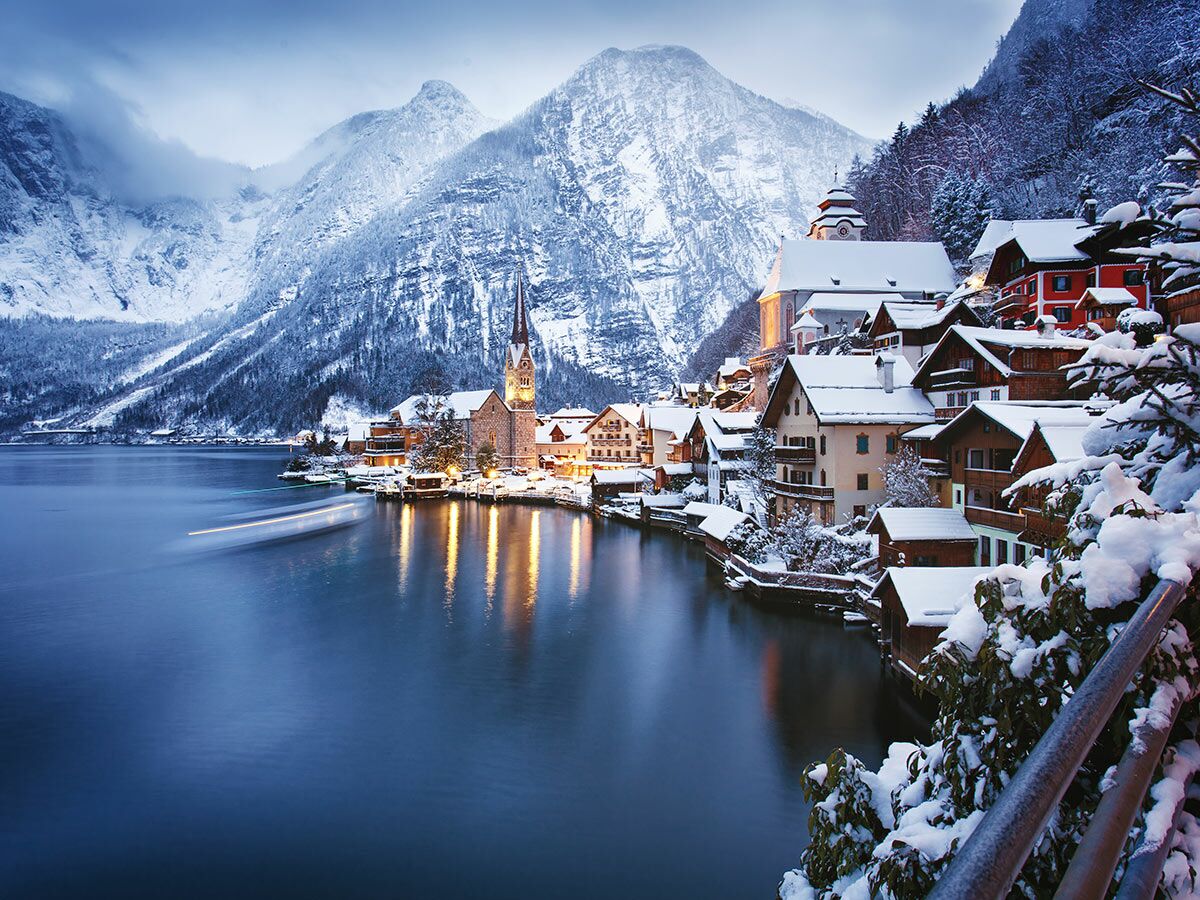 Hallstatt im Winter mit Schnee bedeckten Häusern, See und Alpen. Ein beleuchtetes Dorf mit Kirche spiegelt sich im dunklen Wasser.