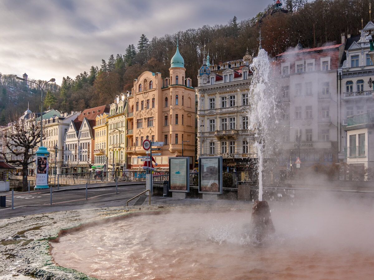 Karlovy Vary: Der berühmte Sprudelkolonnaden-Geysir mit dampfendem Mineralwasserpool und historischen Jugendstilgebäuden im Hintergrund.