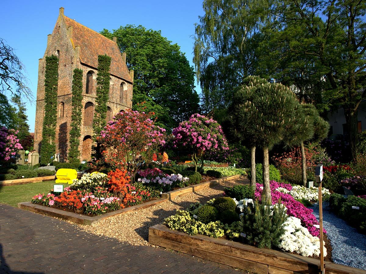 Farbenfroher Garten mit blühenden Rhododendren und Azaleen vor einem historischen Backsteingebäude unter blauem Himmel.