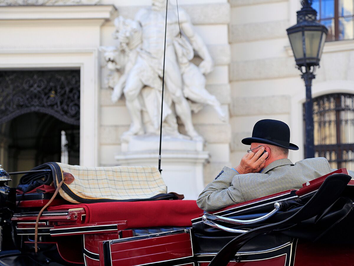 Ein Mann mit Melone sitzt von hinten in einem Fiaker vor historischer Wiener Architektur mit Skulpturen.