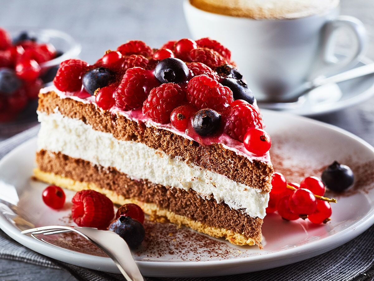 Nahaufnahme eines Stücks Beeren-Sahne-Torte mit Schokoboden, weißer Creme und frischen Beeren auf weißem Teller, Kaffee im Hintergrund.