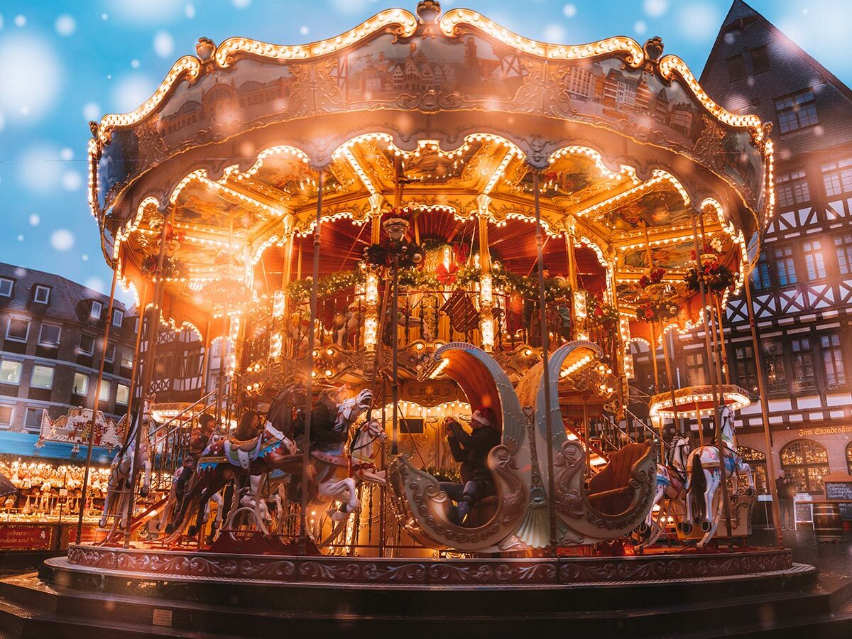 Ein festliches, hell beleuchtetes Karussell auf einem Weihnachtsmarkt bei Schneefall, mit historischen Gebäuden im Hintergrund.