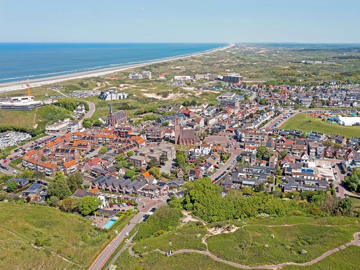 Luftaufnahme einer idyllischen Nordsee-Küstenstadt mit Sandstrand, weiten Dünen und charmanten Häusern unter blauem Himmel.