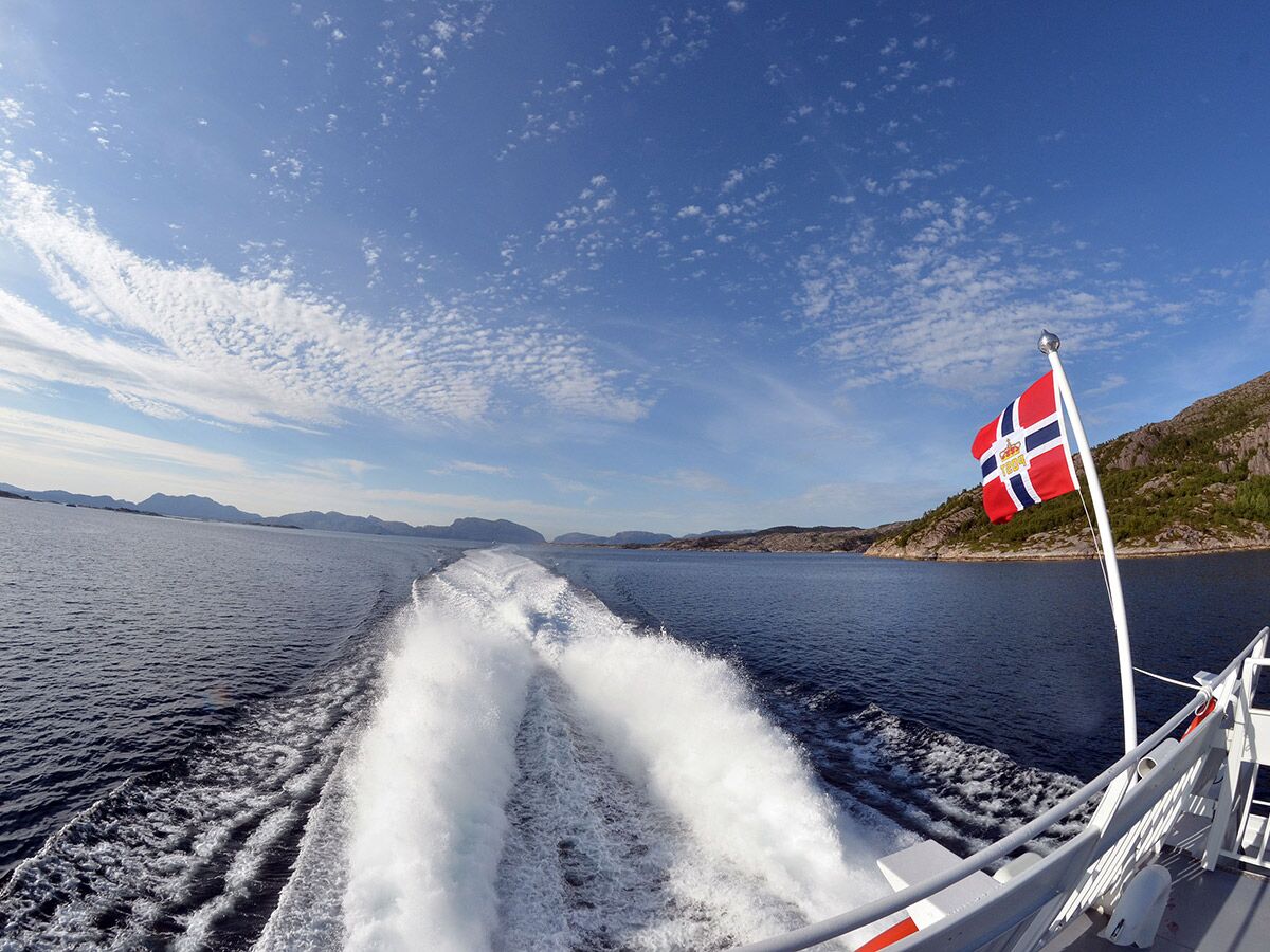 Königliche norwegische Flagge auf einem Schiff, das durch einen sonnigen Fjord fährt und eine große Heckwelle erzeugt.