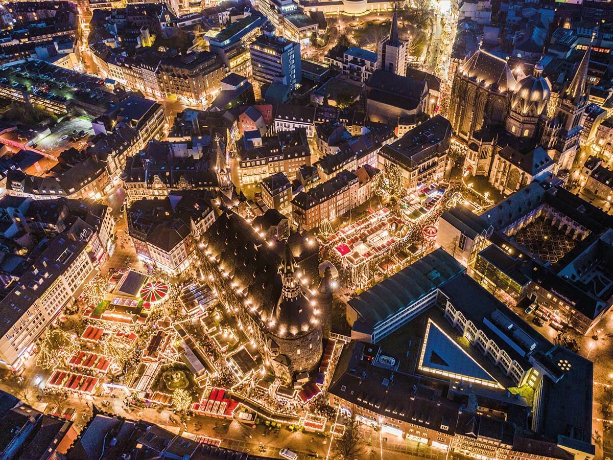 Luftaufnahme des Aachener Weihnachtsmarkts und Doms bei Nacht, mit festlich beleuchteten Ständen und vielen Menschen.