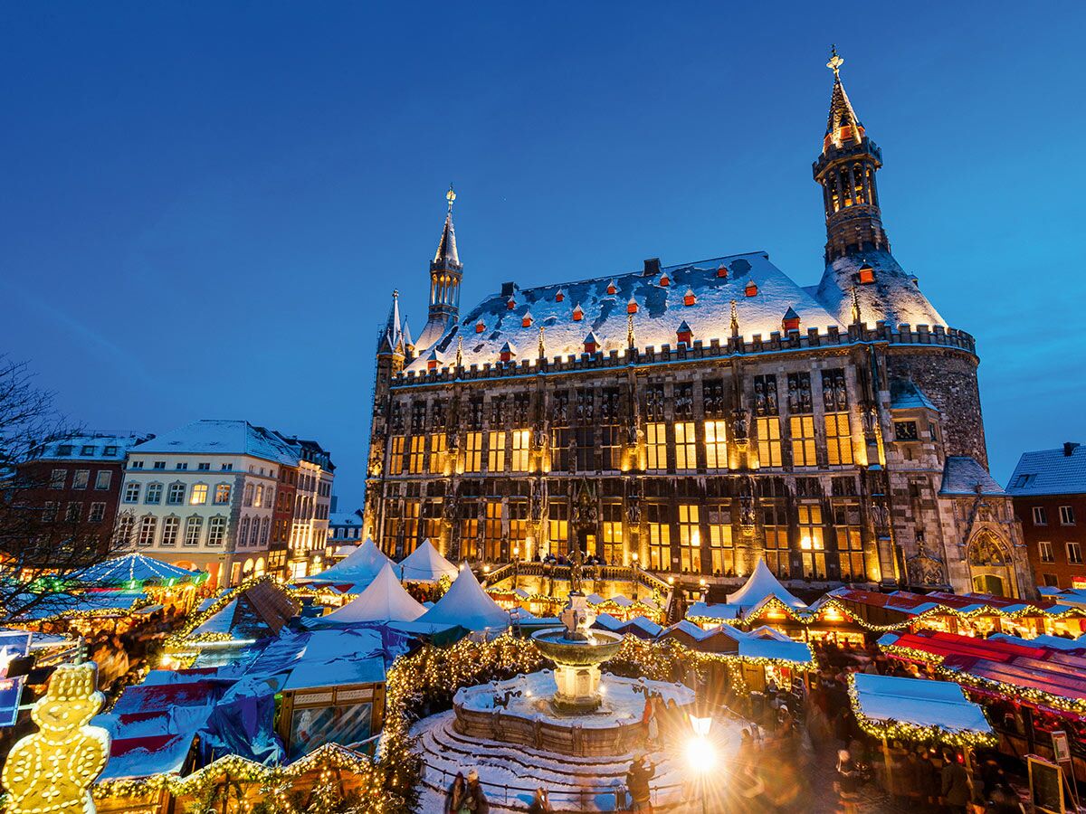 Der festliche Aachener Weihnachtsmarkt im Winter mit beleuchtetem Rathaus, schneebedeckten Ständen und Brunnen bei Nacht.