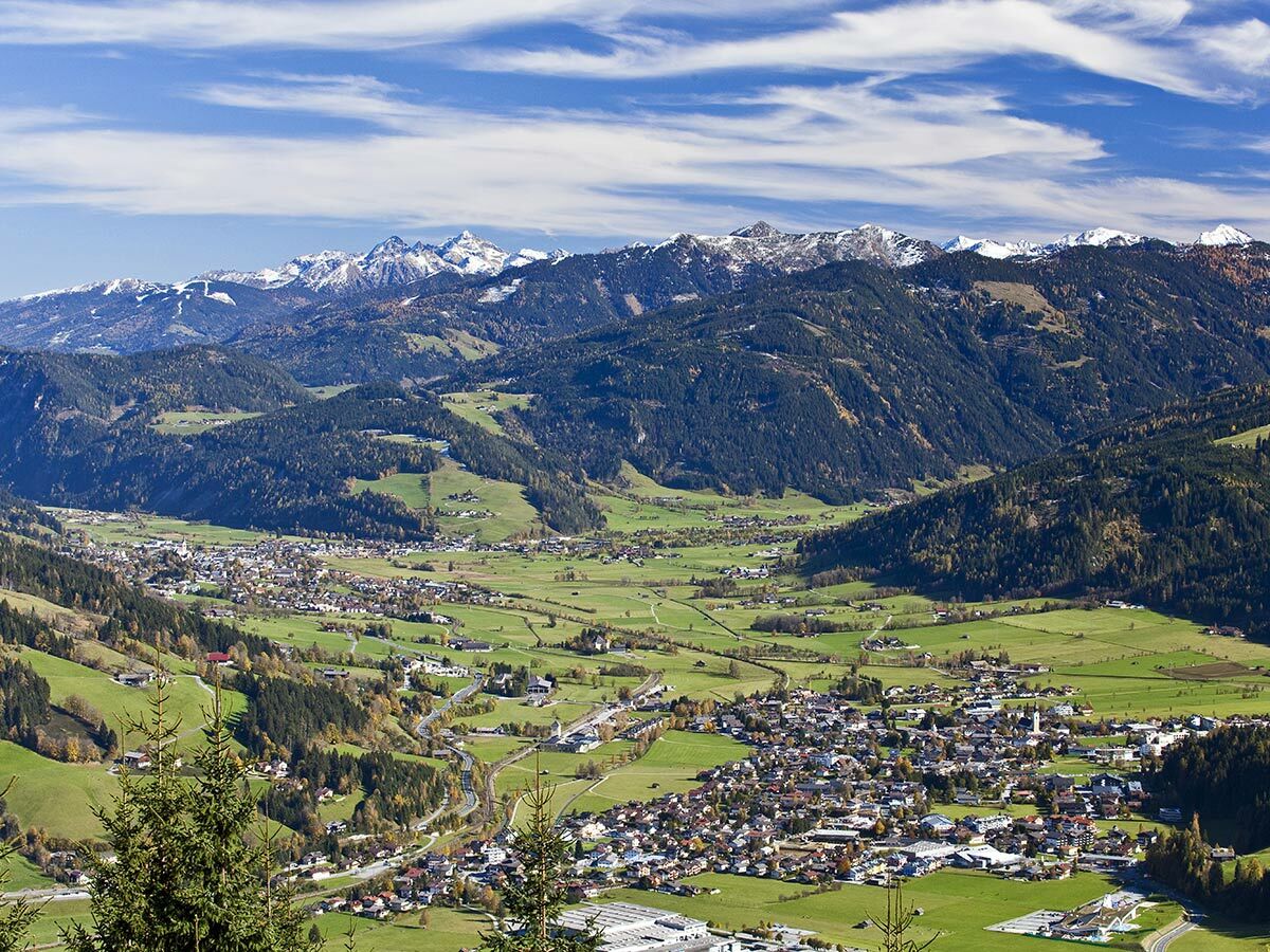 Malerisches Dorf in einem grünen Tal, umgeben von Wäldern und schneebedeckten Alpenbergen unter blauem Himmel.