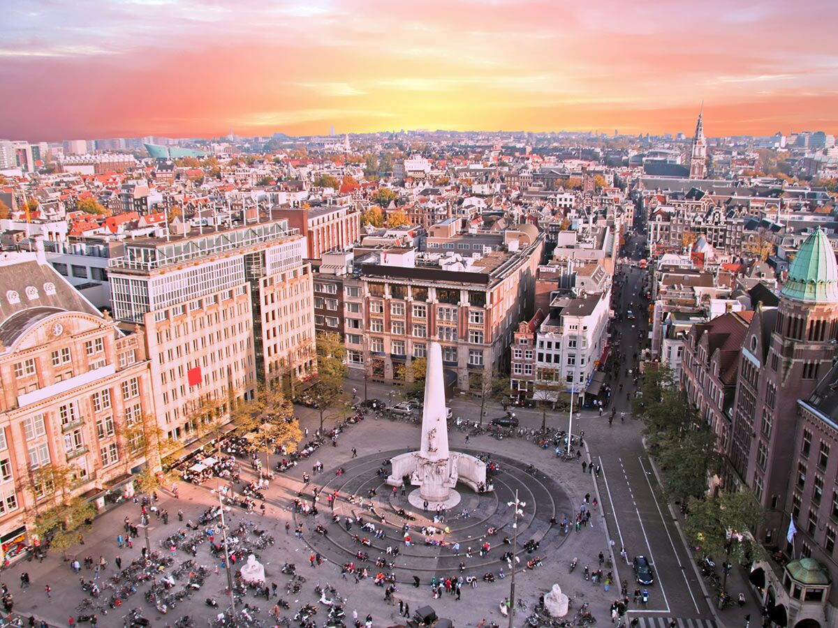 Luftaufnahme des Dam Platzes in Amsterdam bei Sonnenuntergang mit dem Nationalmonument, historischen Gebäuden und vielen Menschen.