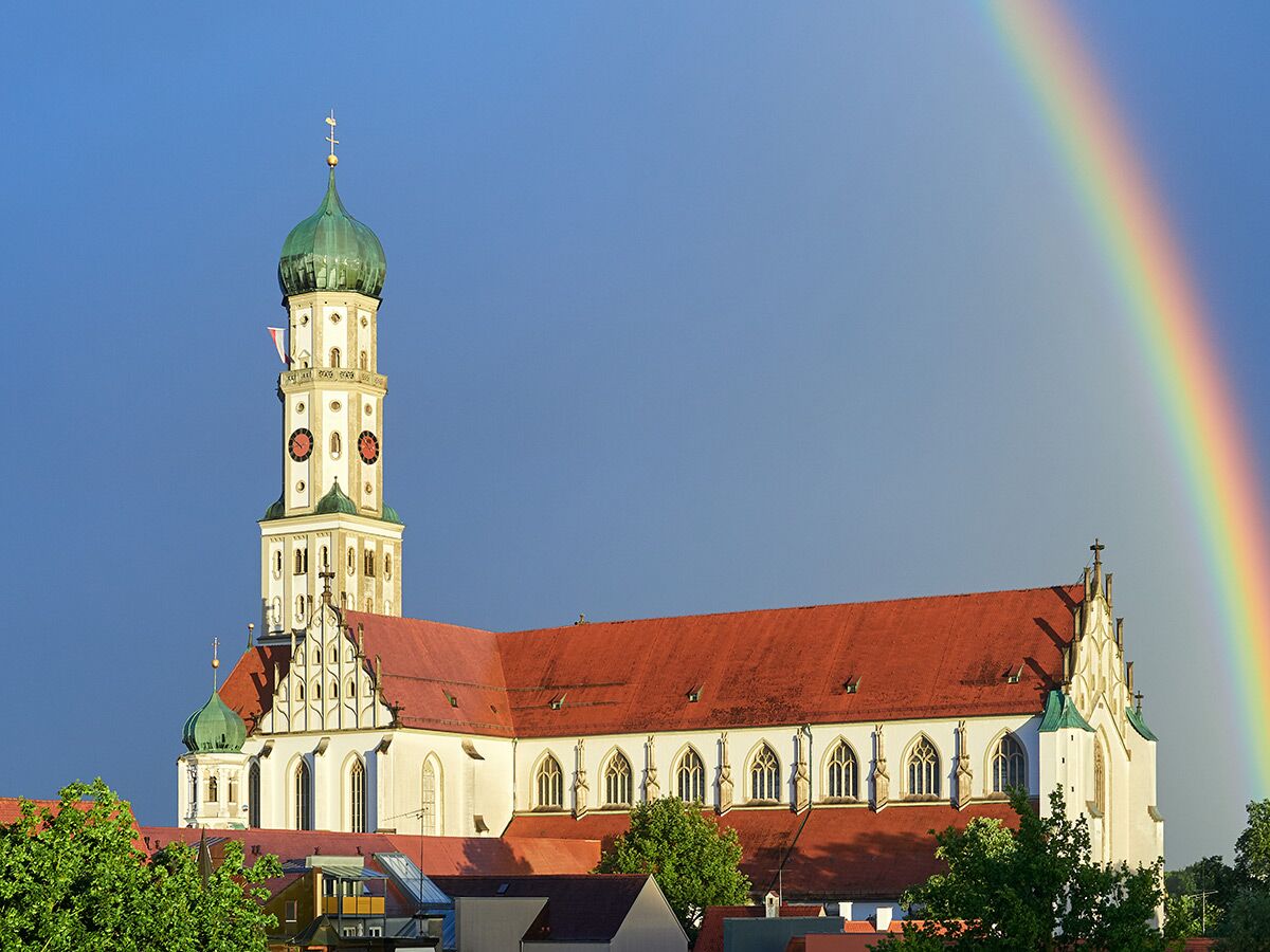 Historische Basilika St. Ulrich und Afra in Augsburg mit markantem Zwiebelturm, rotem Dach und leuchtendem Regenbogen am blauen Himmel.