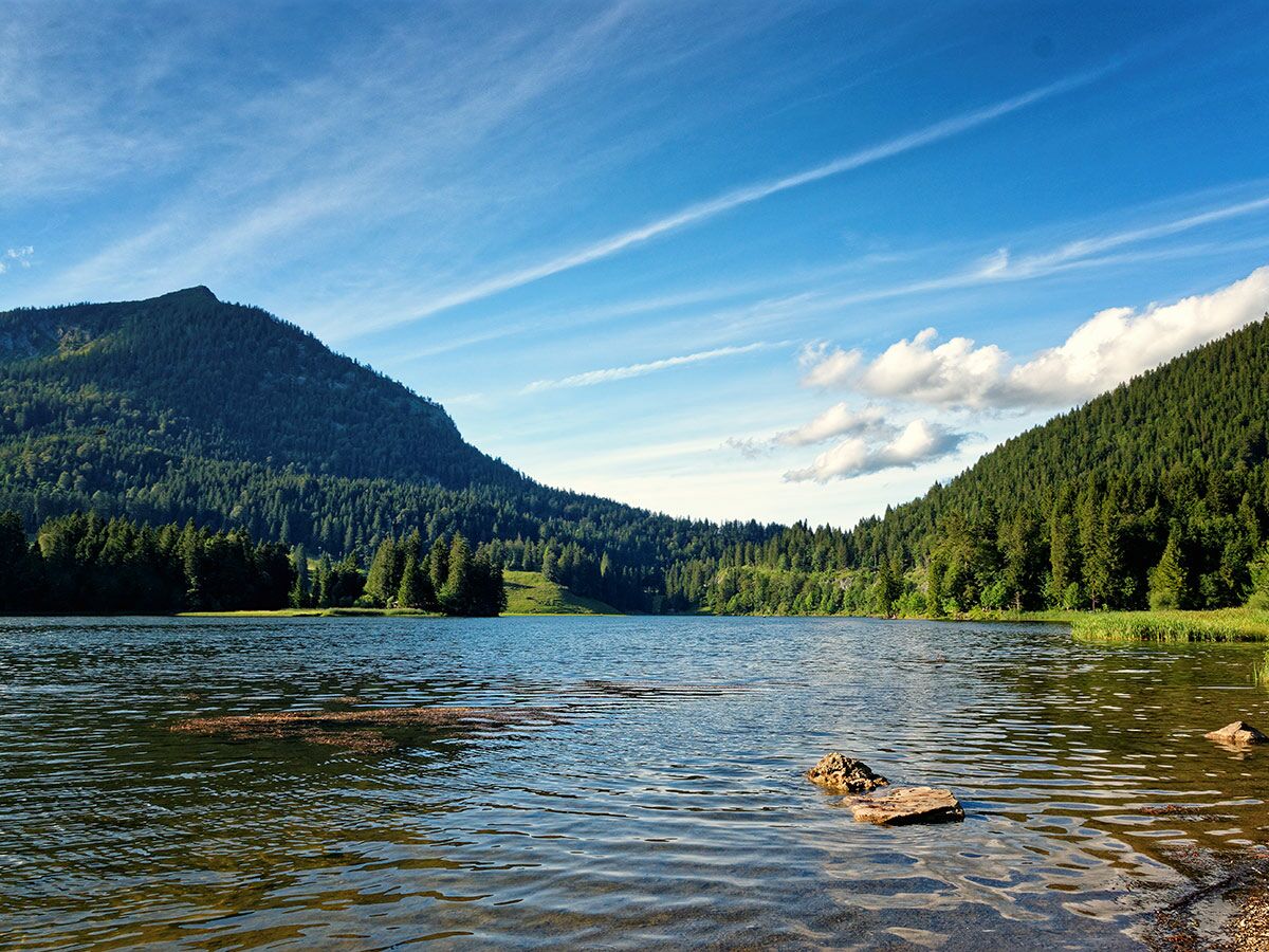Ruhiger Bergsee umgeben von dichten grünen Wäldern und majestätischen Bergen unter einem strahlend blauen Himmel mit Wolken.