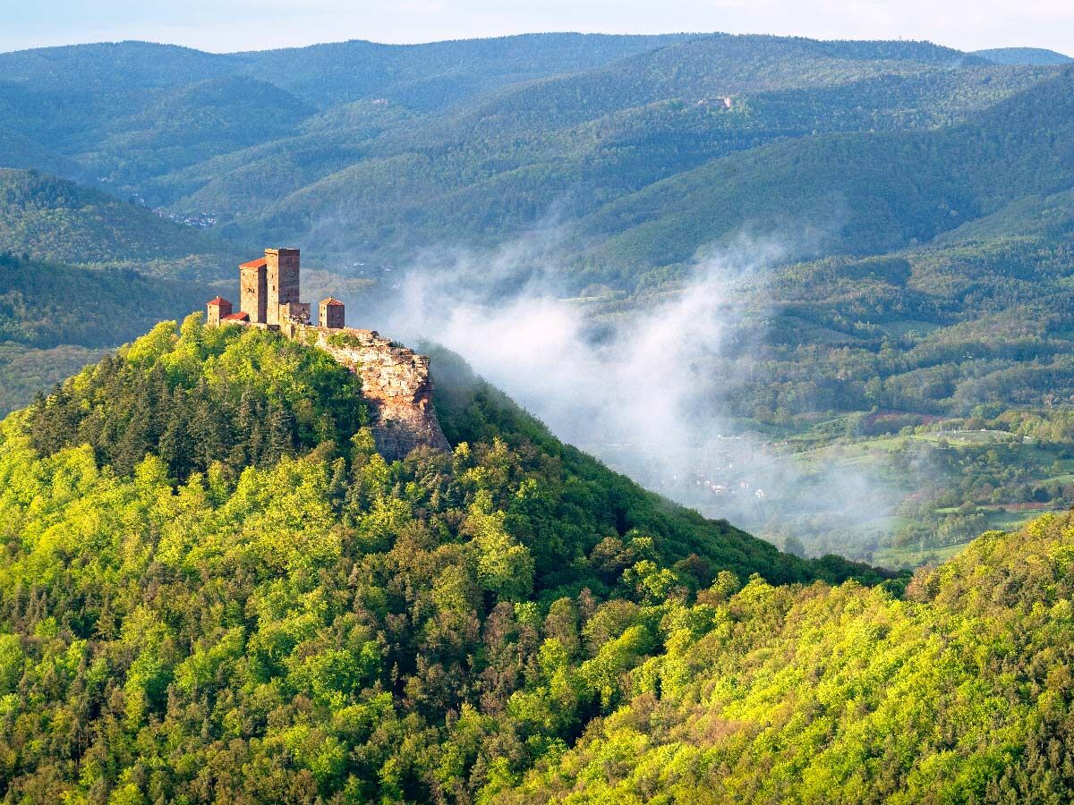 Historische Burg auf grün bewaldetem Hügel, umgeben von sanften Bergen und mystischem Nebel in einer weiten Landschaft.