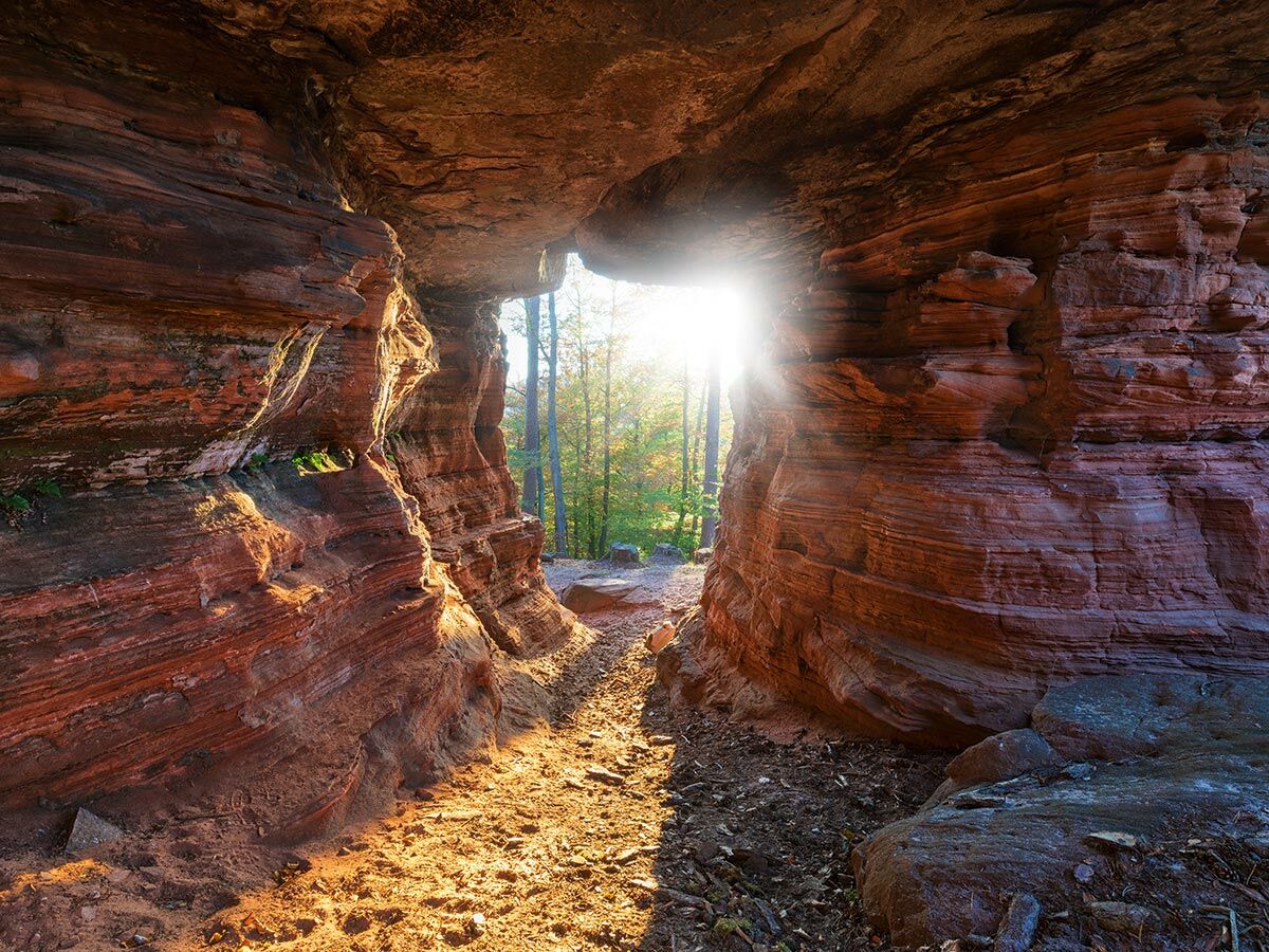Sonnenlicht strahlt durch eine rote Sandstein-Felsformation, die einen natürlichen Höhleneingang zu einem Waldpfad bildet.