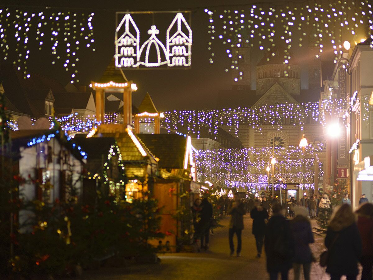 Festlicher Weihnachtsmarkt bei Nacht, geschmückt mit unzähligen Lichtern, Menschen schlendern vor Domkulisse und Marktständen.