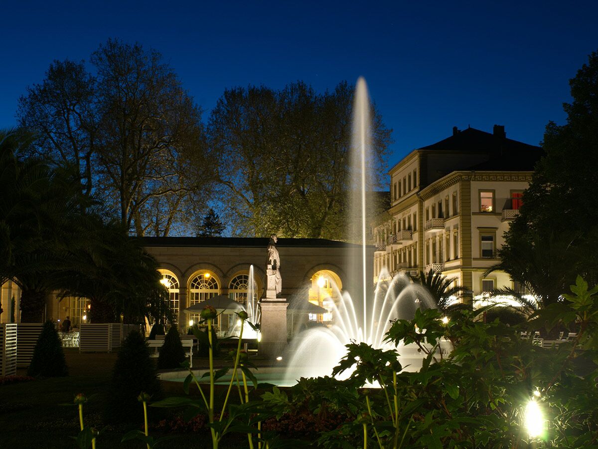 Nachtaufnahme eines beleuchteten Parks mit Springbrunnen, einem eleganten Gebäude mit Balkonen und hohen Bäumen unter blauem Himmel.