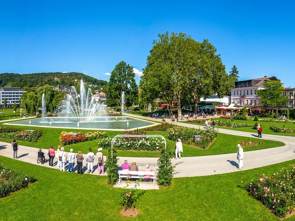Sonnenverwöhnter Kurpark mit großem Springbrunnen, blühenden Rosenbeeten und Menschen, die spazieren gehen. Im Hintergrund historische Gebäude.