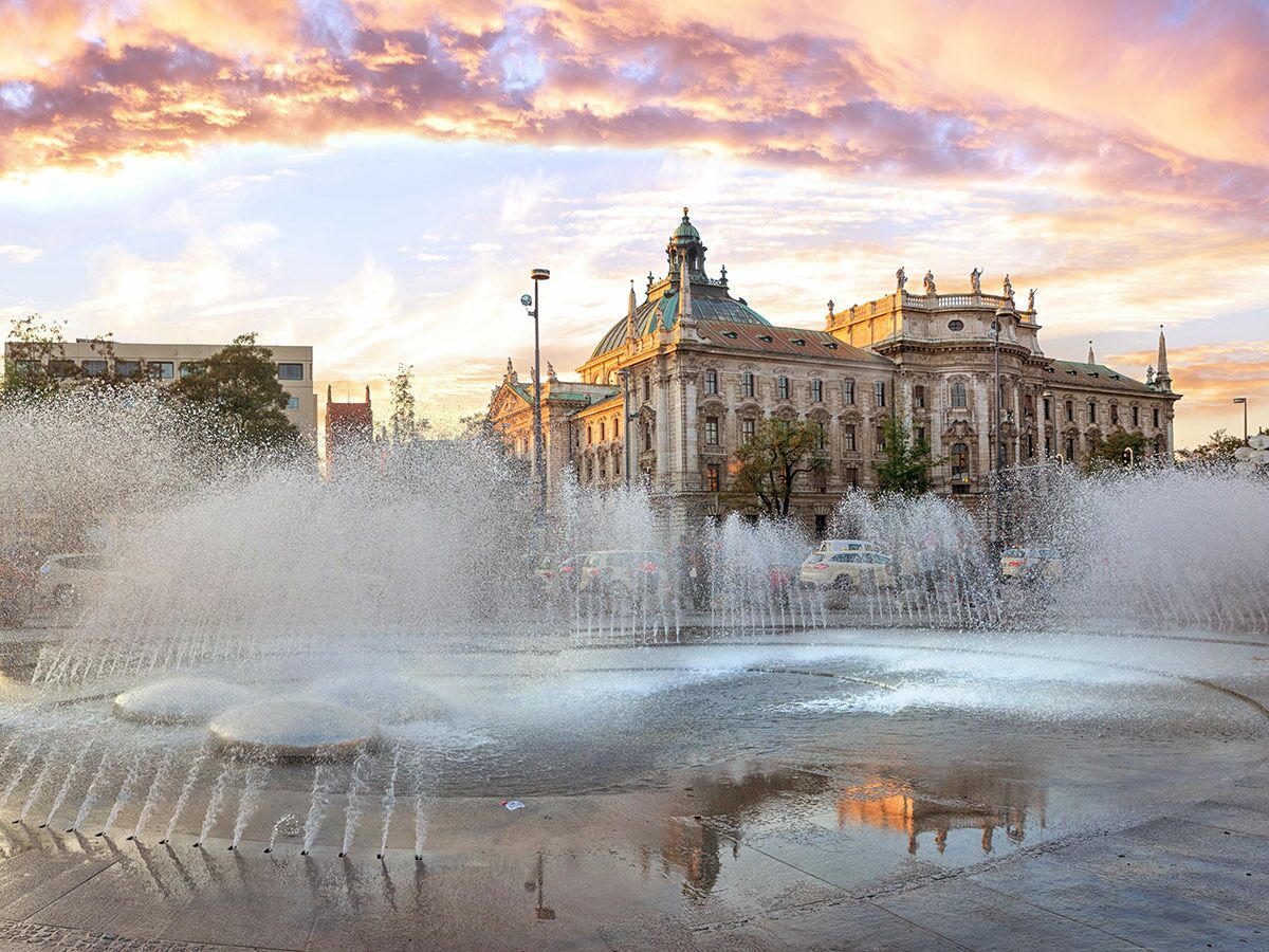 Historischer Justizpalast in München bei Sonnenuntergang, beleuchtet von warmem Licht, mit Springbrunnen im Vordergrund und reflektierendem Wasser.