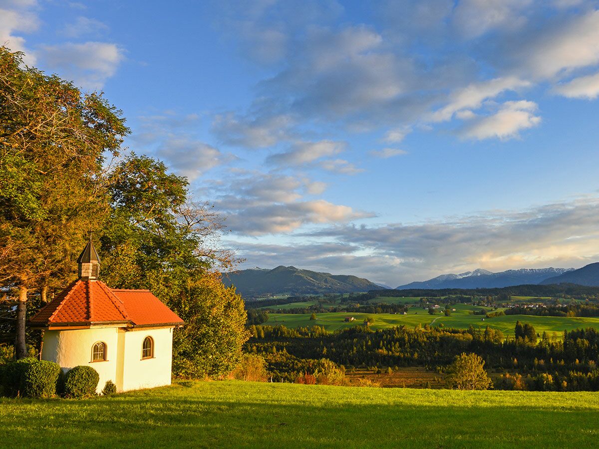 Kleine weiße Kapelle mit rotem Dach zwischen grünen Wiesen und Bäumen, majestätische Berge im Hintergrund bei goldenem Licht.