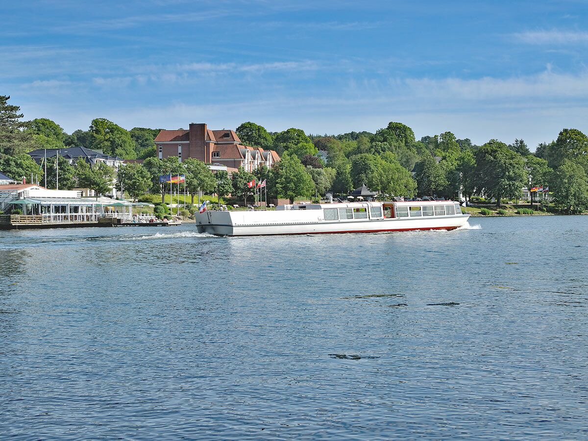 Weißes Passagierschiff mit rotem Streifen fährt über einen See, am grünen Ufer stehen Gebäude und wehen Flaggen unter blauem Himmel.