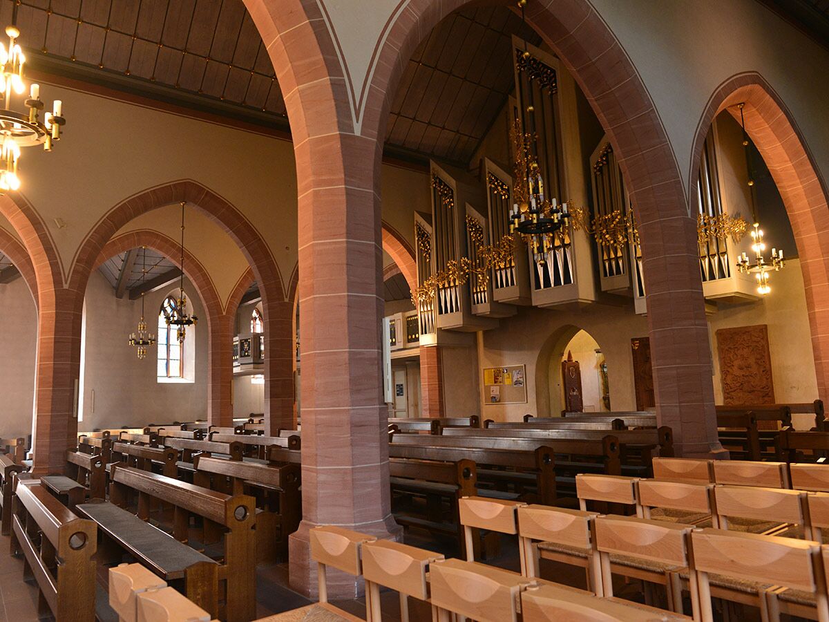 Innenansicht einer Kirche mit hohen roten Steinbögen, Reihen von Holzbänken und einer prächtigen Pfeifenorgel.