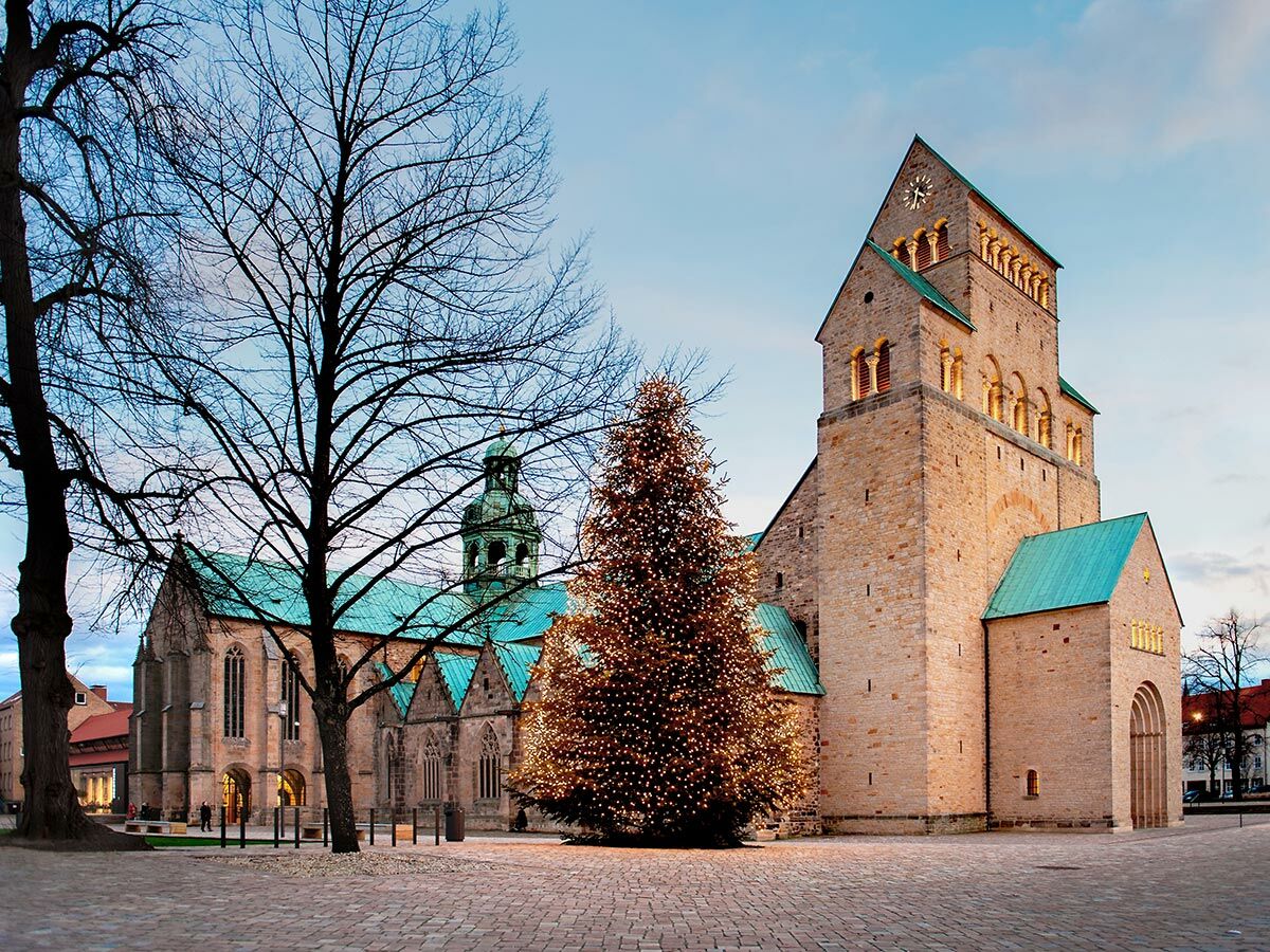 Der Hildesheimer Dom mit grünem Dach und beleuchtetem Weihnachtsbaum auf einem gepflasterten Platz in der Dämmerung.